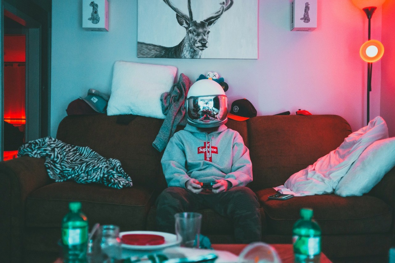 Junge Person mit Astronautenhelm auf einem Sofa, umgeben von Chaos, spielt Videospiel – Sinnbild für Eskapismus, Identitätssuche und persönliche Entwicklung.