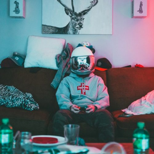Junge Person mit Astronautenhelm auf einem Sofa, umgeben von Chaos, spielt Videospiel – Sinnbild für Eskapismus, Identitätssuche und persönliche Entwicklung.